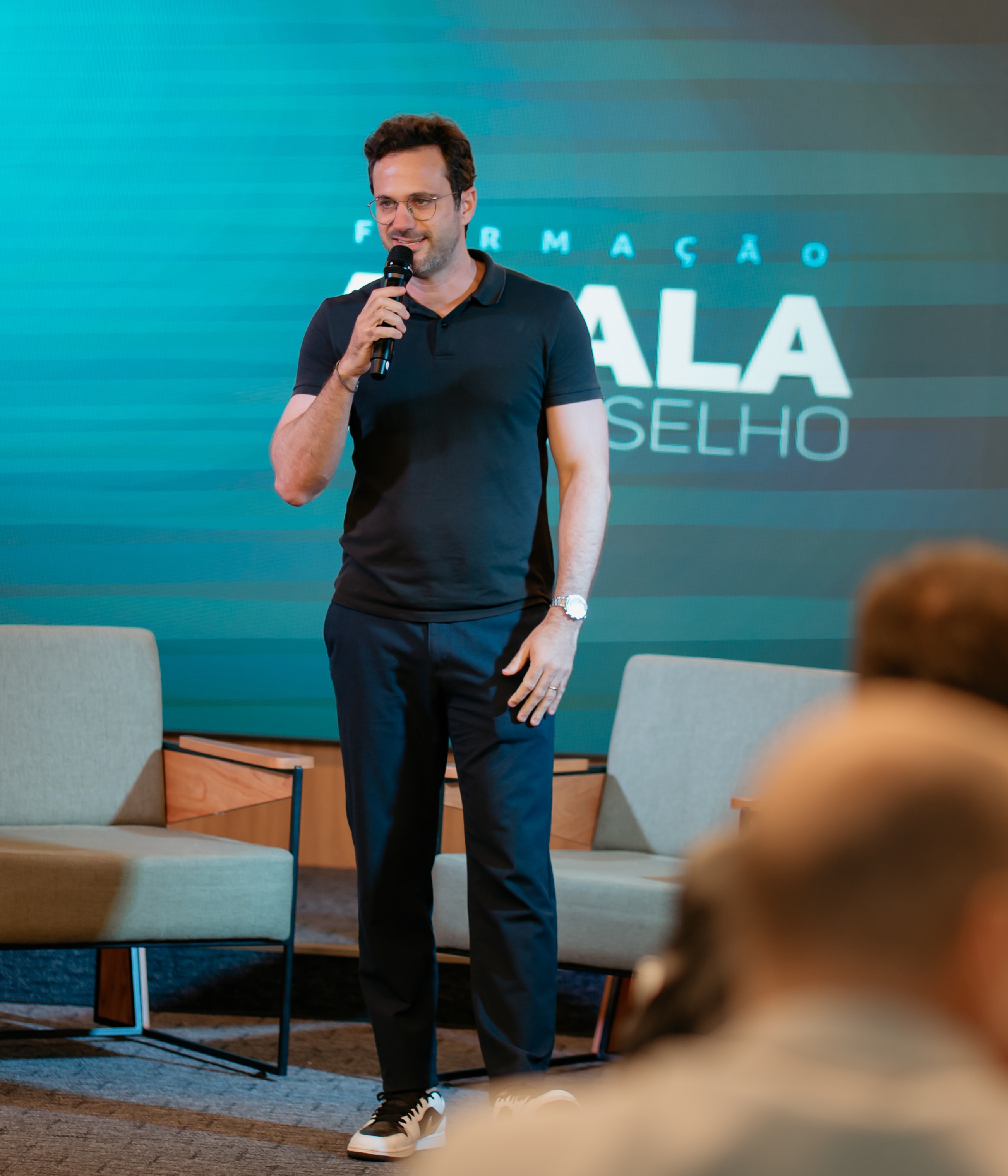 Thiago Lago no palco da formação A Sala, microfone na mão, painel com o nome do evento ao fundo.